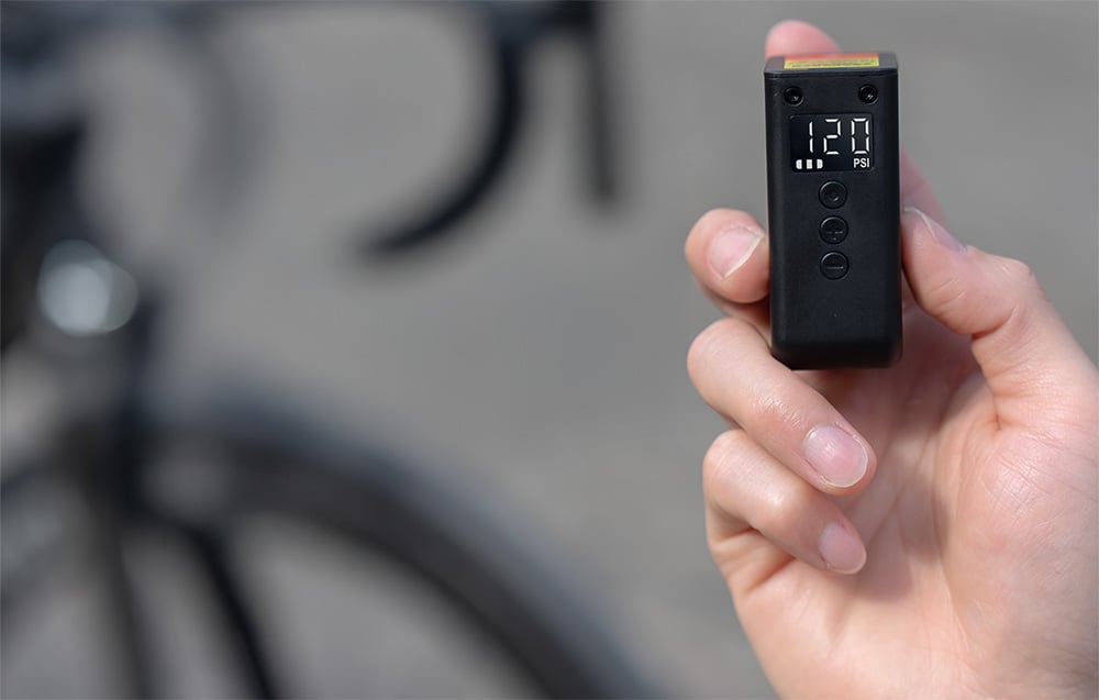 Dłoń rowerzysty trzymająca ultralekką pompkę akumulatorową Cycplus na tle szprych koła rowerowego.